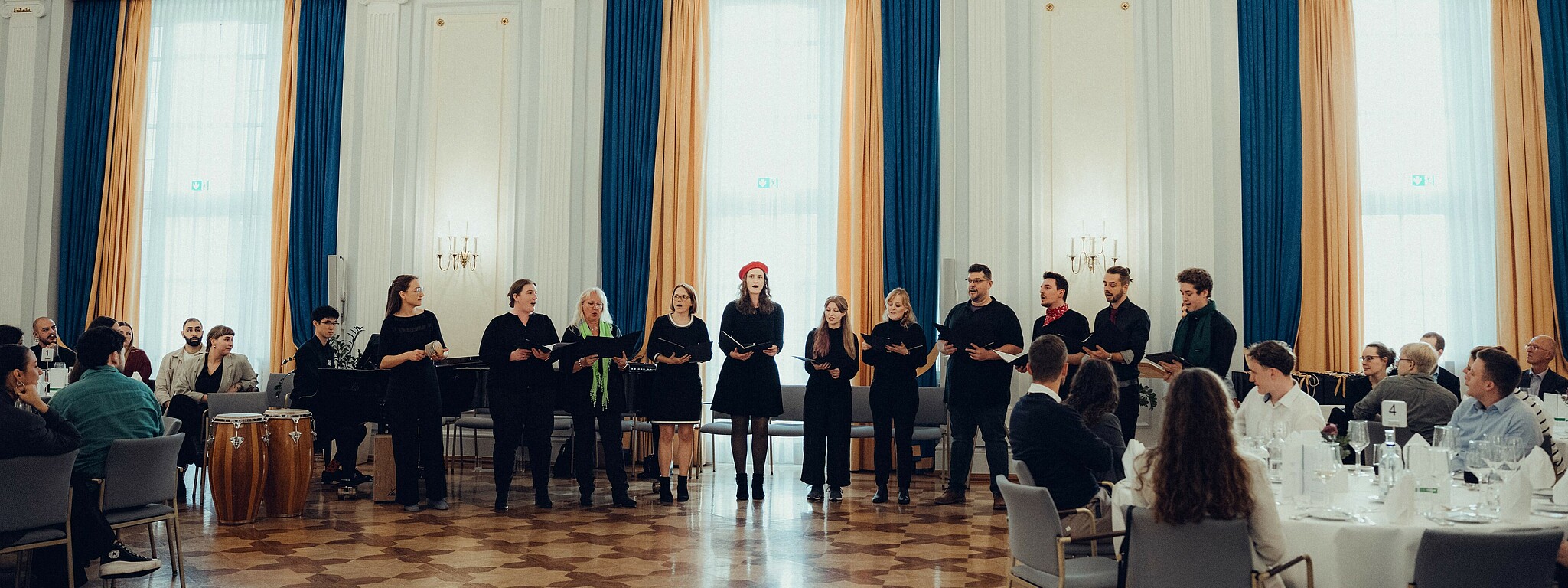 Der Hochschulchor steht in einer Reihe vor einem sitzenden Publikum in einem festlichen Raum mit hohen Fenstern und blauen sowie goldenen Vorhängen und singt gemeinsam.