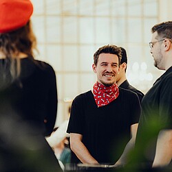 Nahaufnahme eines männlichen Chorsängers, der lächelt und ein rotes Halstuch trägt, während er mit anderen Chormitgliedern interagiert.
