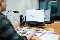 Ein Monitor zeigt das Logo der Hochschule für Wirtschaft und Gesellschaft Ludwigshafen, während eine Person vor dem Bildschirm sitzt und Materialien auf dem Tisch betrachtet. Verschiedene farbige Karten und Euroscheine sind auf dem Tisch verteilt.